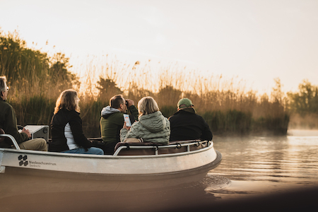 Bezoekers in een klein bootje turen in de verte. Het is nog heel vroeg, de lucht is nog lichtoranje en er hangt een beetje mist boven het water.